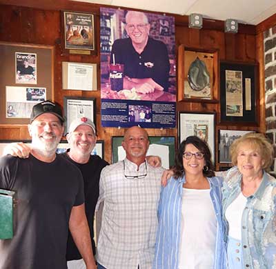 A group of smiling people stand in front of a wall full of photos and press clippings
